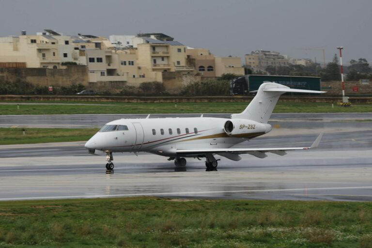 A private jet taxis on a wet runway at an airport with buildings in the background.