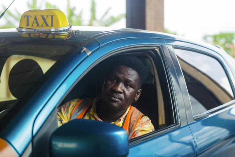 A taxi driver sitting in a blue car, wearing an orange vest, waiting for passengers.